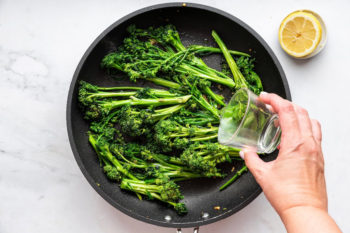 A woman's hand pours a small amount of water into a pan of broccolini.