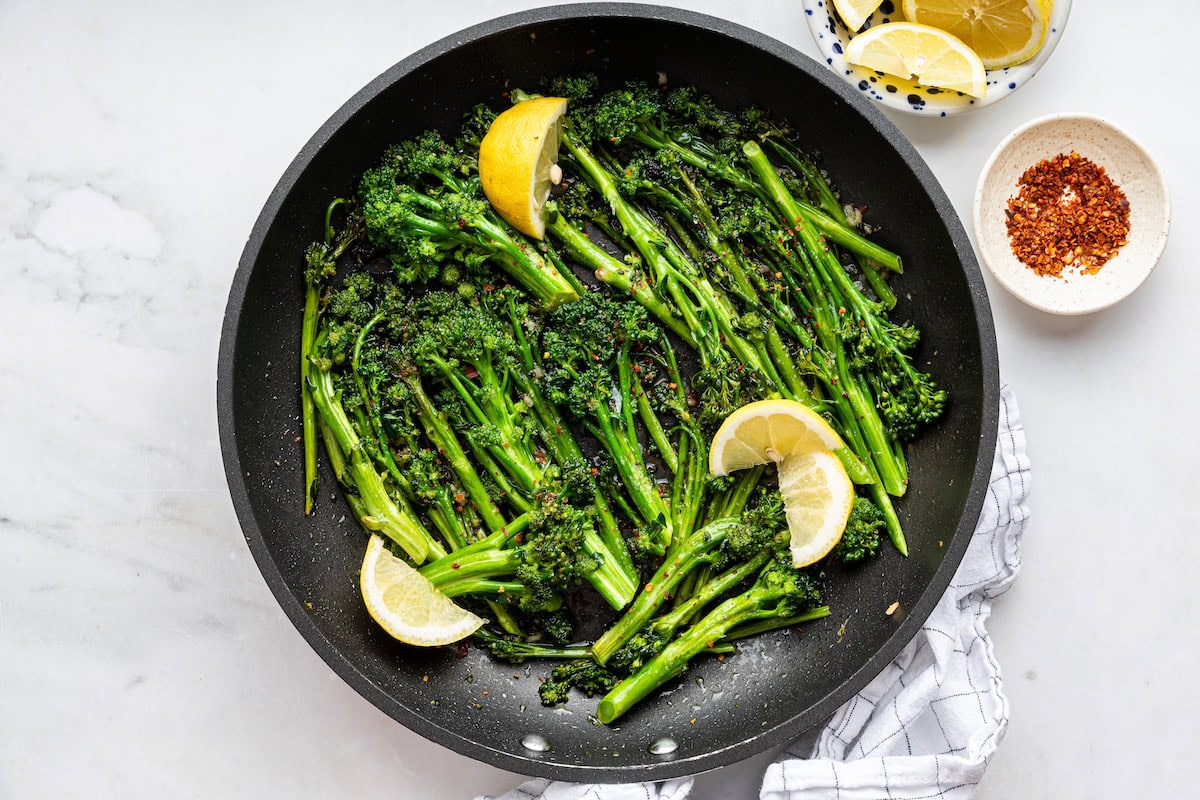 A non-stick cooking pan with sauteed broccolini and fresh lemon wedges.