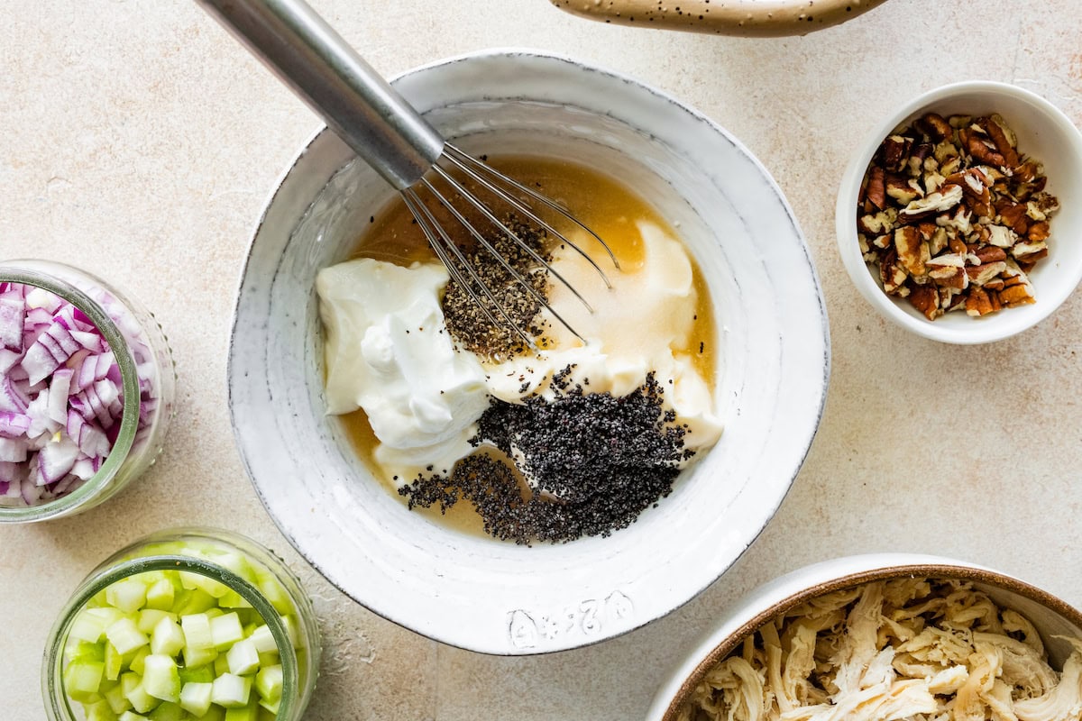 A small bowl with a metal whisk that is going to whisk greek yogurt, mayo, vinegar, poppy seeds, and black pepper to make a dressing.
