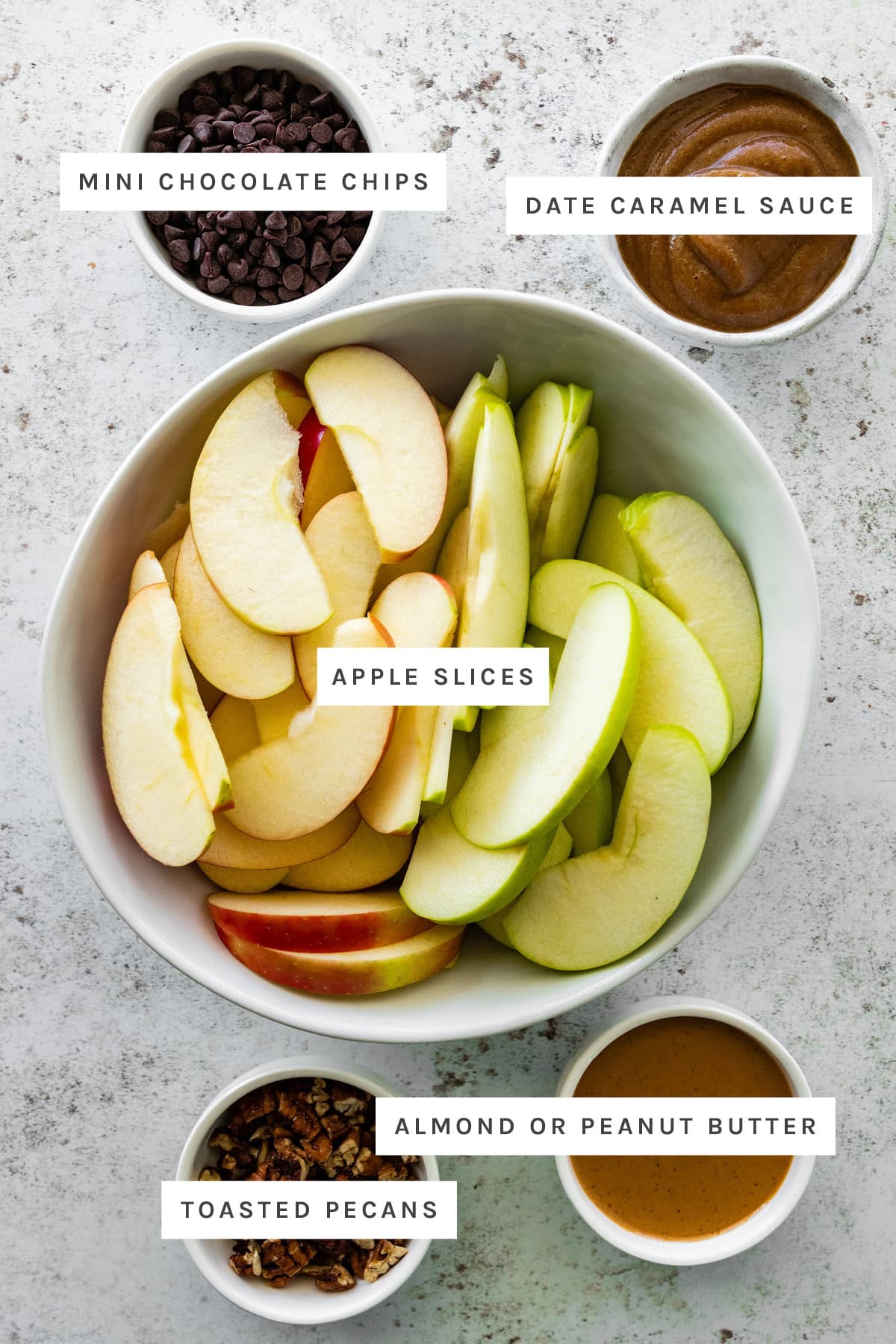 Ingredients measured out to make Healthy Apple Nachos with Date Caramel Sauce: mini chocolate chips, date caramel sauce, apple slices, toasted pecans and almond or peanut butter.