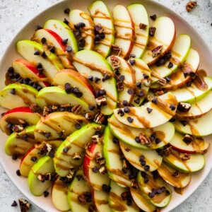 A large plate with a display of apple slices that are topped with a drizzle of date caramel, mini chocolate chips, and crushed pecans.