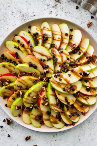 A large plate with a display of apple slices that are topped with a drizzle of date caramel, mini chocolate chips, and crushed pecans.