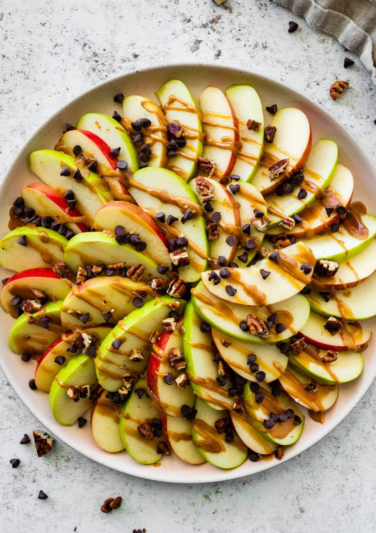 A large plate with a display of apple slices that are topped with a drizzle of date caramel, mini chocolate chips, and crushed pecans.