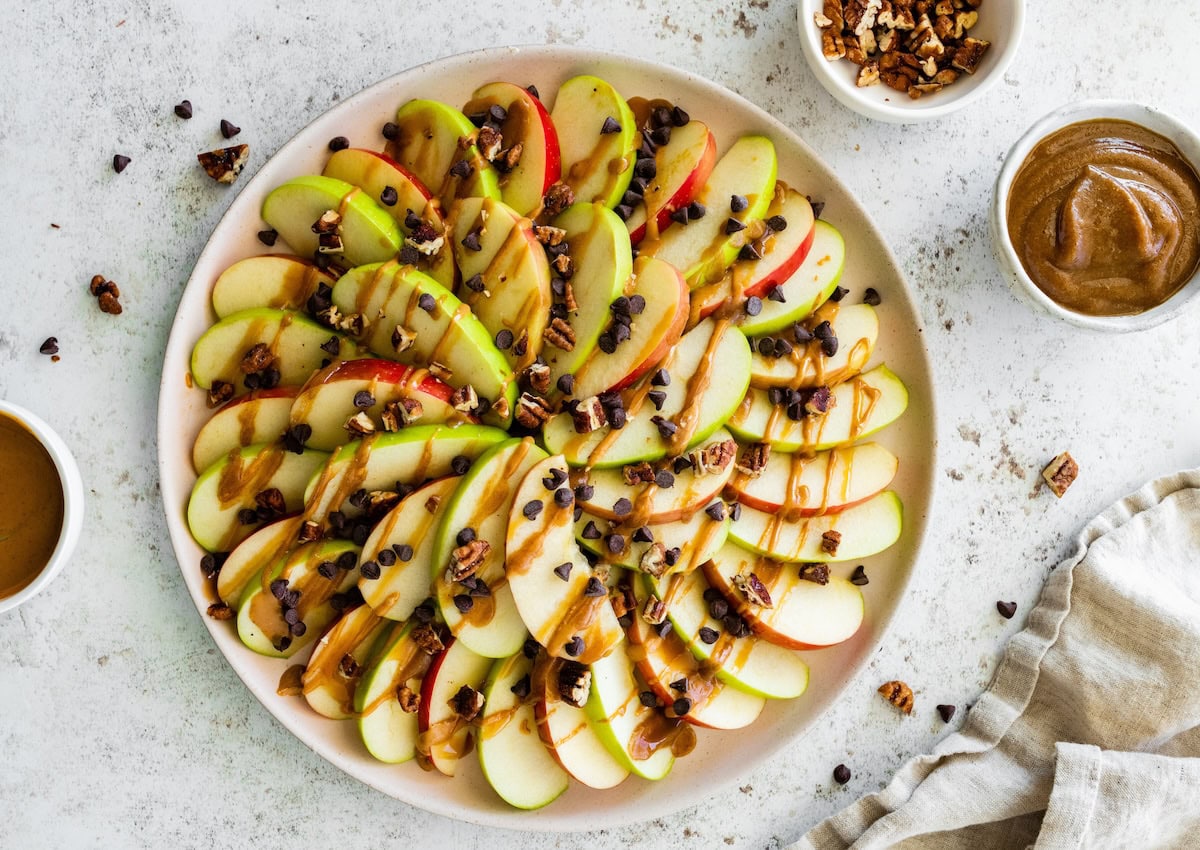 A large plate with apple slices that are topped with a drizzle of date caramel, chocolate chips, and pecans.
