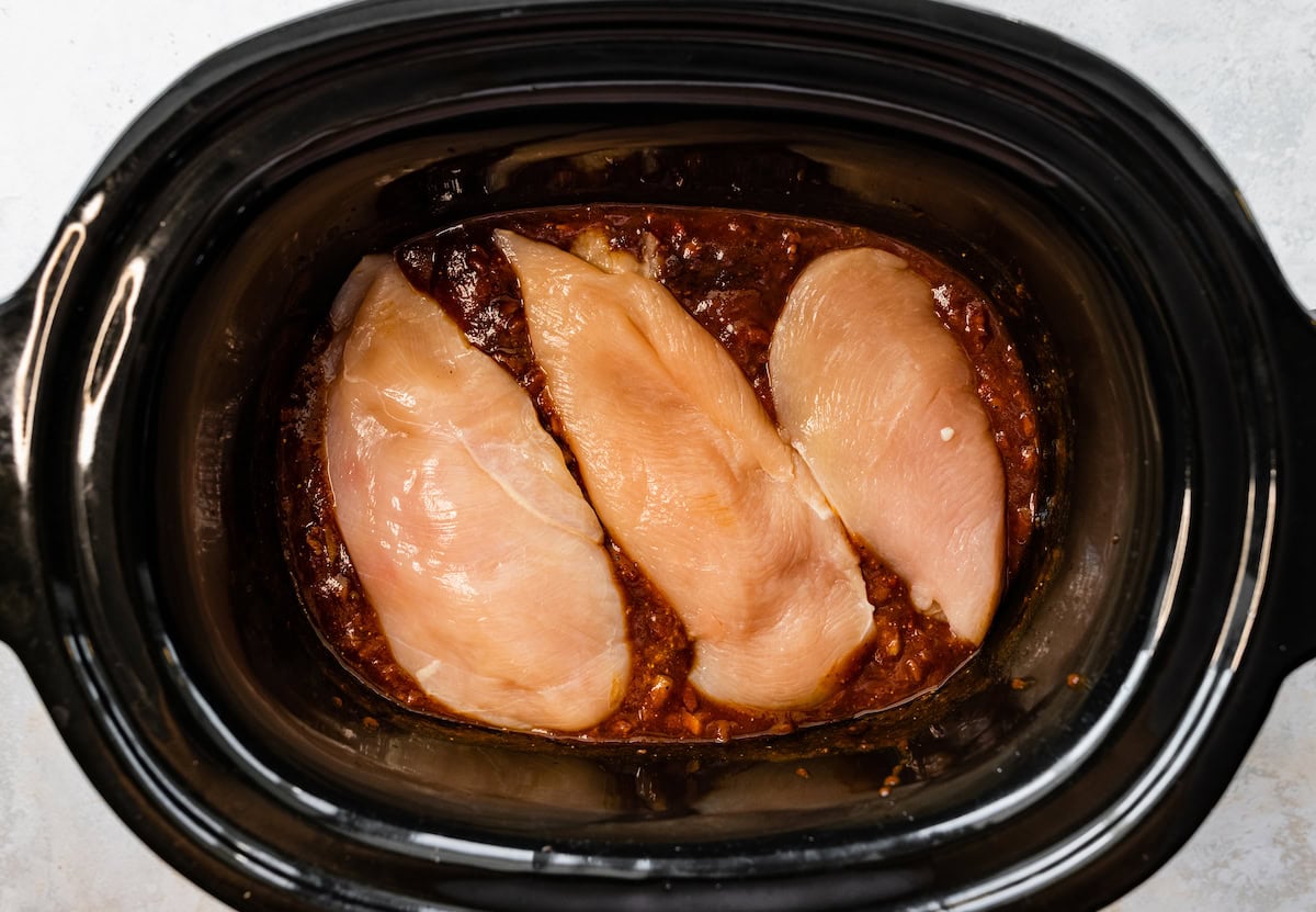 Three raw chicken breasts in a slow cooker with bbq sauce and spices.