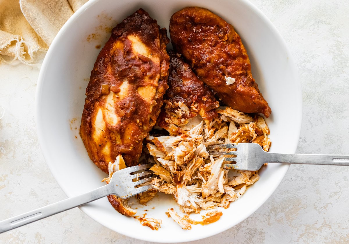 BBQ chicken breasts are being shredded in a bowl with two forks.