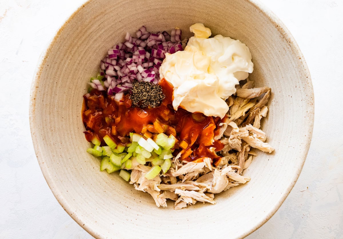 A large mixing bowl of shredded chicken, celery, buffalo sauce, mayo, yogurt, red onion, and spices.