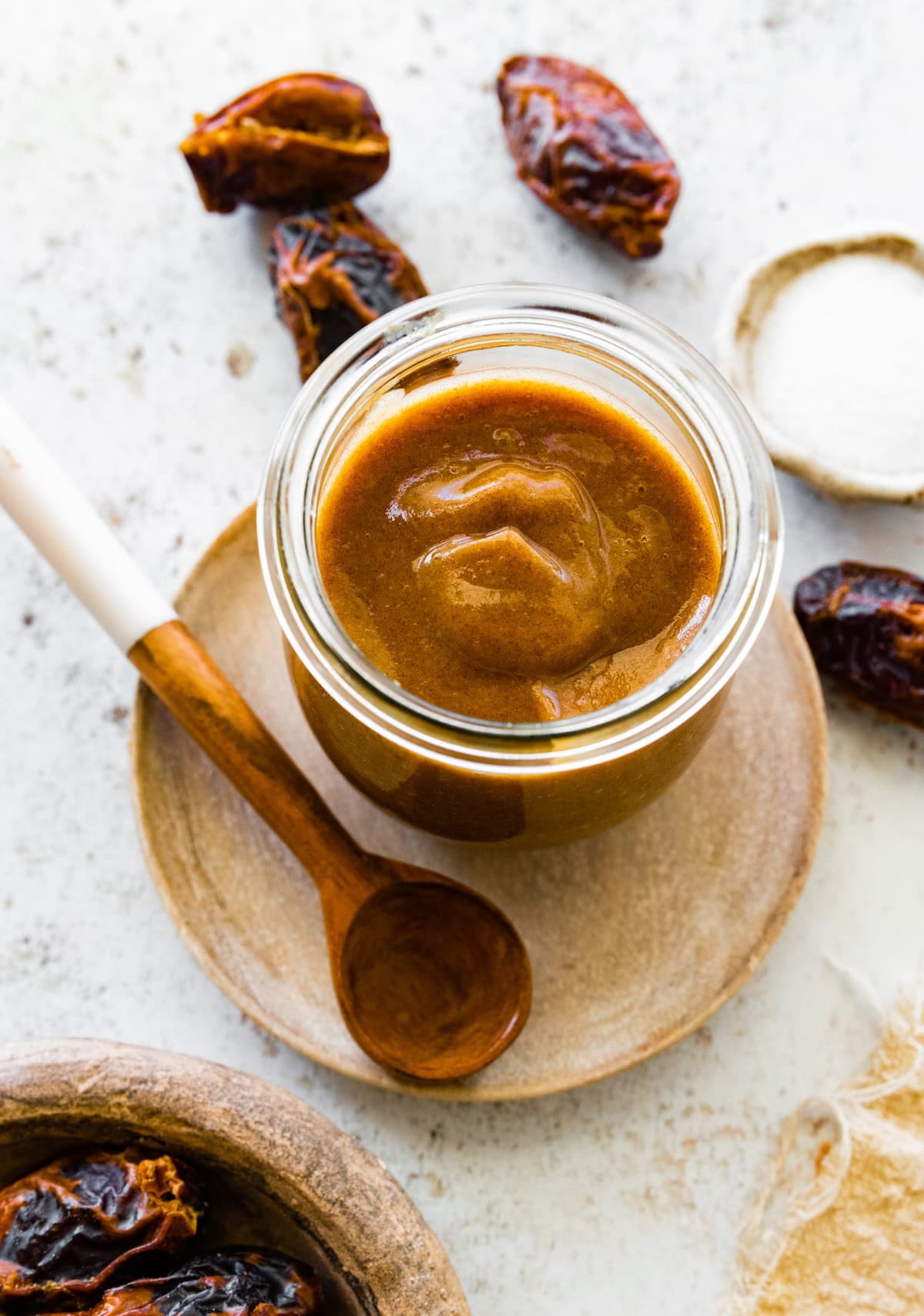 A glass jar of date caramel near a wooden spoon and Medjool dates.