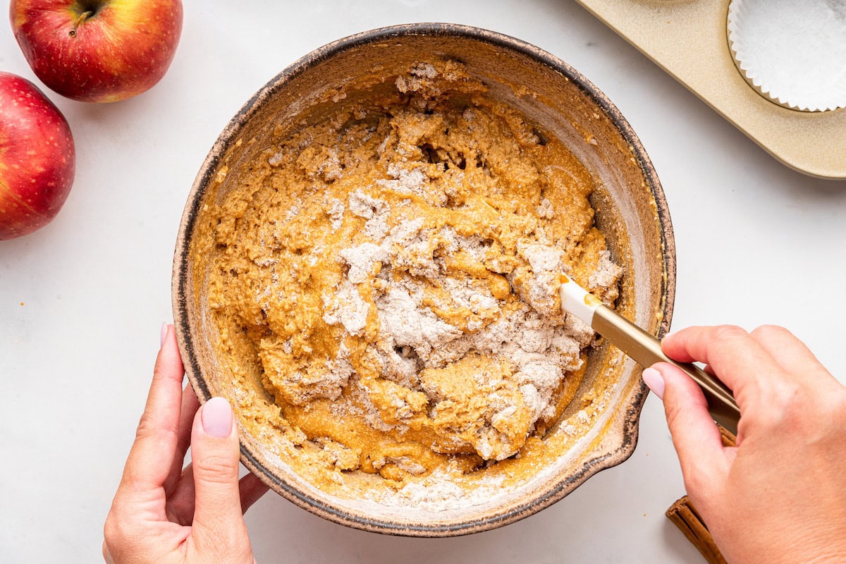 A woman's hand uses a silicone spatula to combine dry and wet ingredients used for apple muffins in a large mixing bowl.