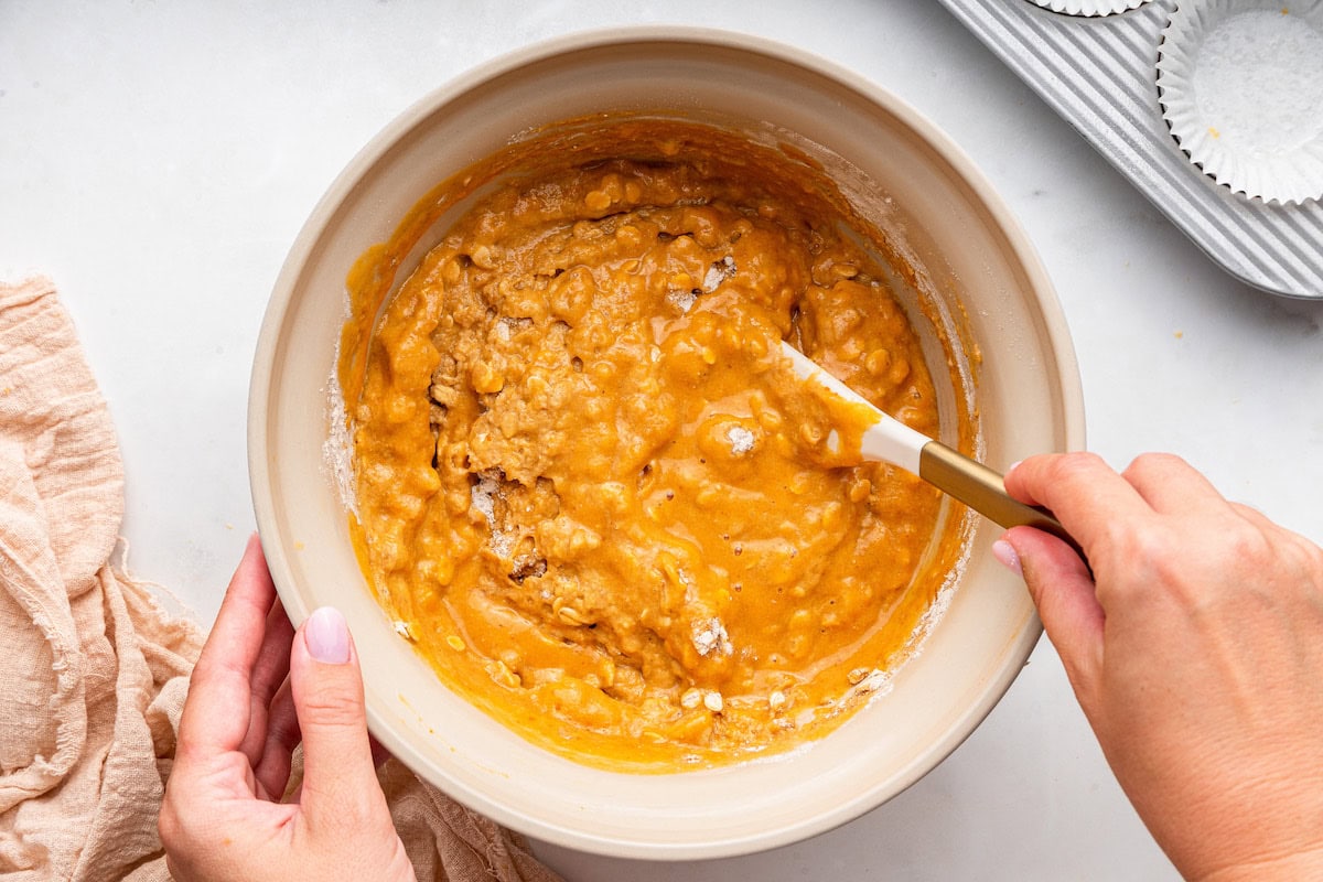 A woman's hand uses a silicone spatula to fold the wet and dry ingredients for pumpkin muffins together.