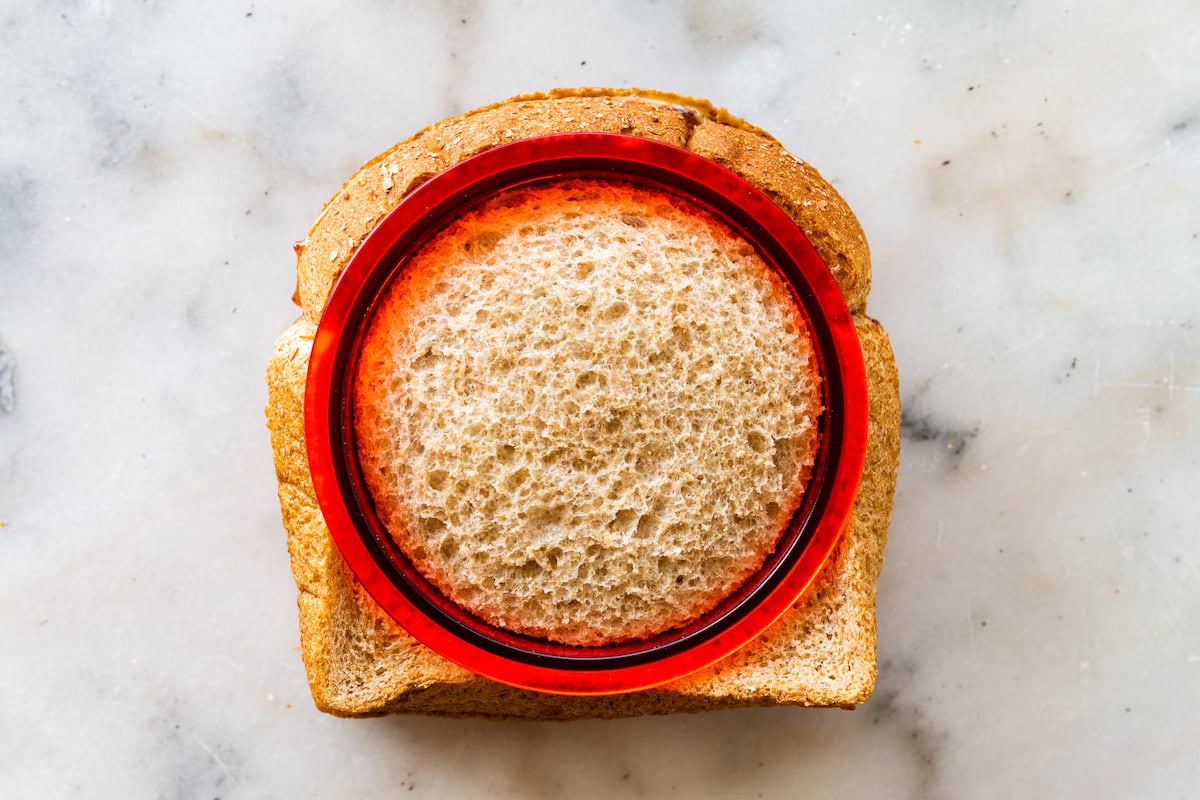 A round sandwich sealer on top of a peanut butter and jelly sandwich, is used to cut the crust off.