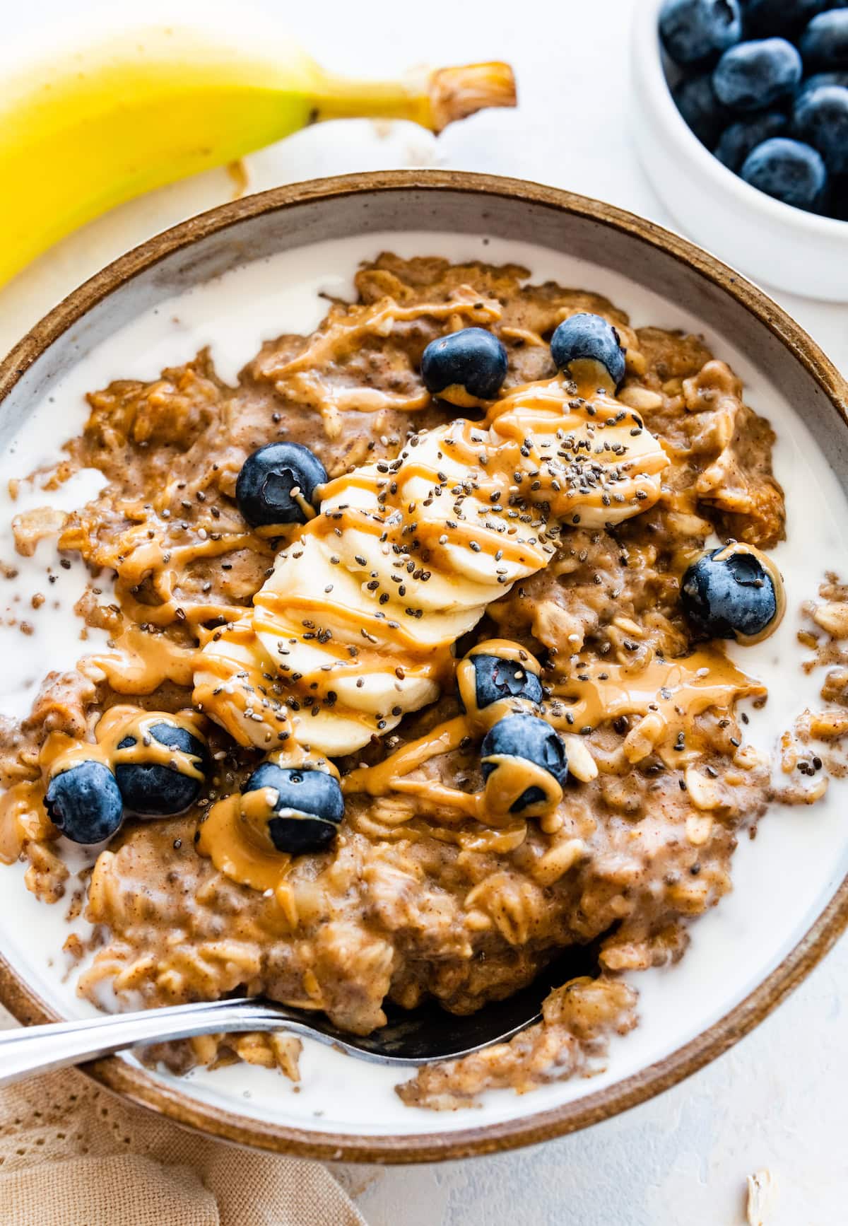 A serving of peanut butter oatmeal in a bowl with milk and topped with banana slices, blueberries, chia seeds, and a drizzle of peanut butter.