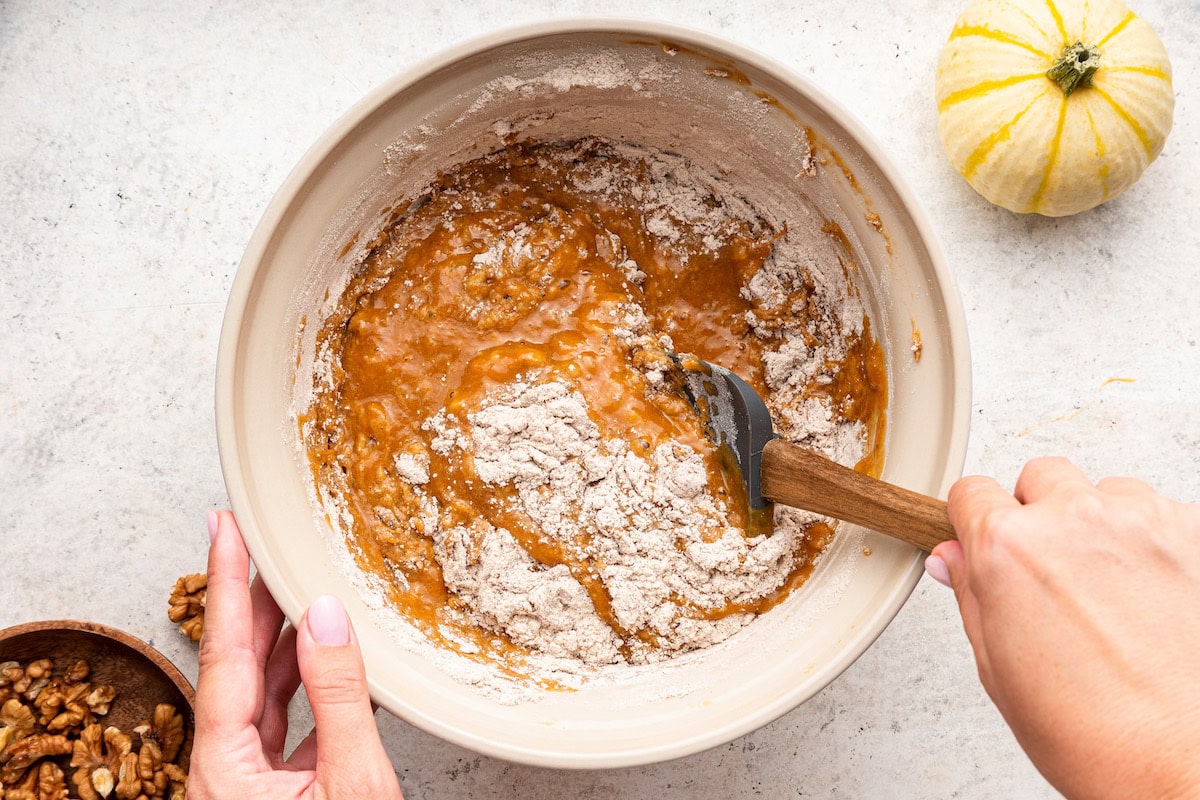 A woman's hand uses a silicone spatula to combine the wet and dry ingredients for a pumpkin banana bread.