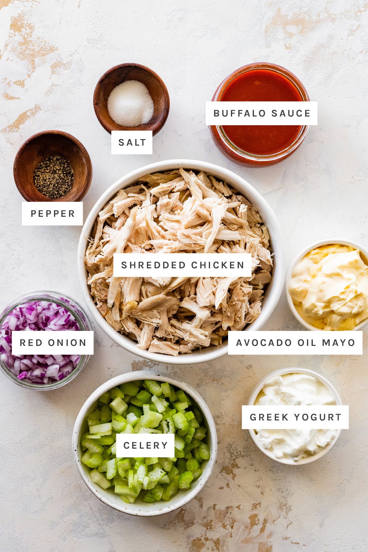 Ingredients measured out to make Buffalo Chicken Salad: salt, pepper, buffalo sauce, shredded chicken, avocado oil mayo, red onion, celery and Greek yogurt.