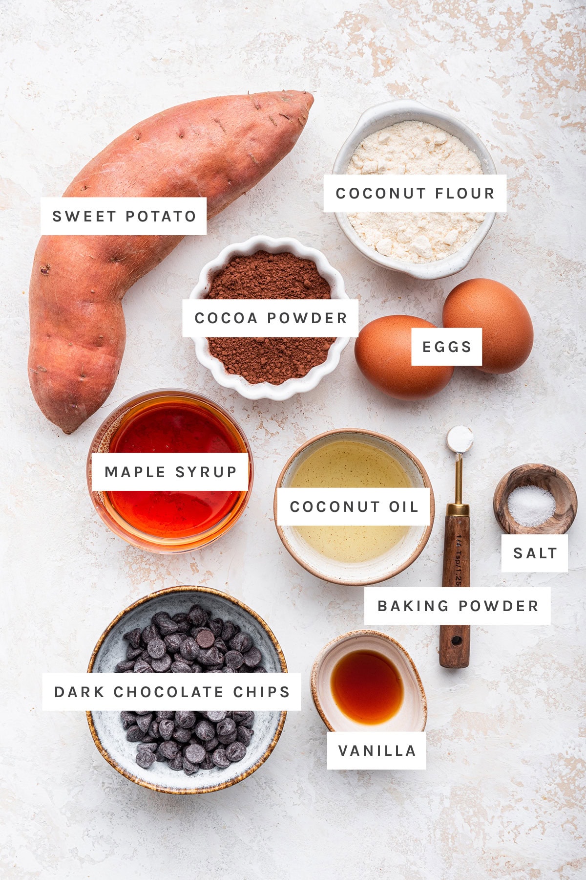 Ingredients measured out to make Sweet Potato Brownies: sweet potato, coconut flour, cocoa powder, eggs, maple syrup, coconut oil, baking powder, salt, vanilla and dark chocolate chips.