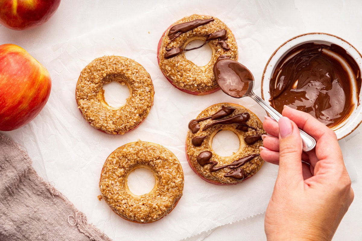 A woman's hand uses a spoon to drizzle chocolate over apple slices with almond butter, coconut, and maple syrup on top.