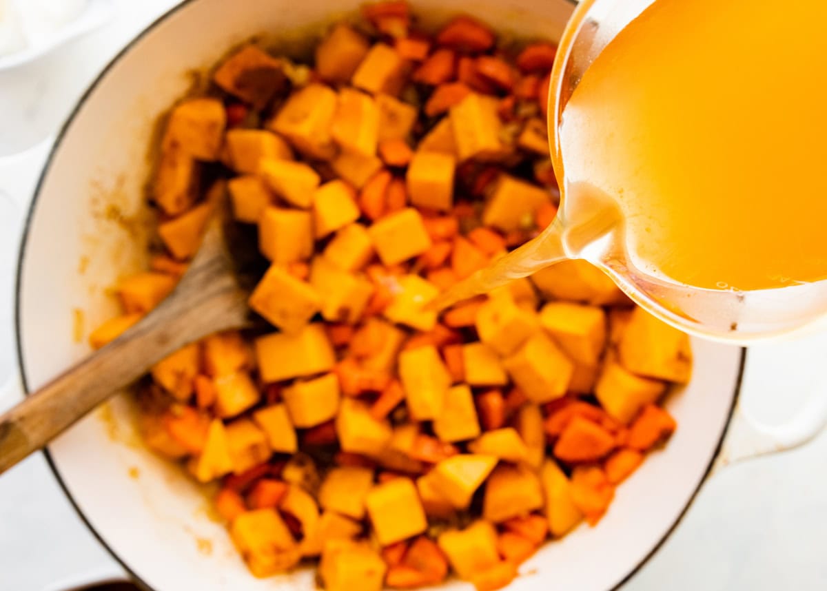 Broth being poured into a large pot of squash, carrot, onions, ginger, garlic, and other warm spices.
