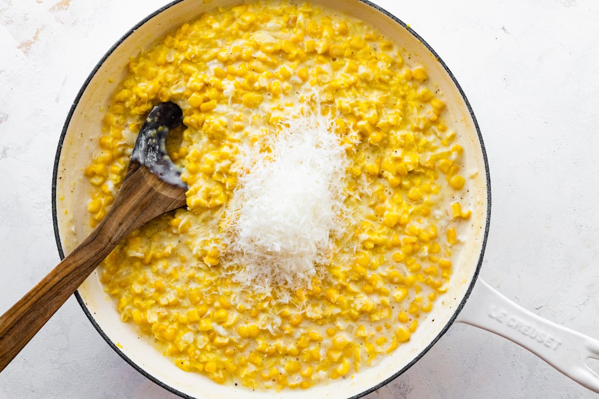 Freshly grated parmesan being mixed into the creamed corn mixture in a white skillet with a wooden spoon.
