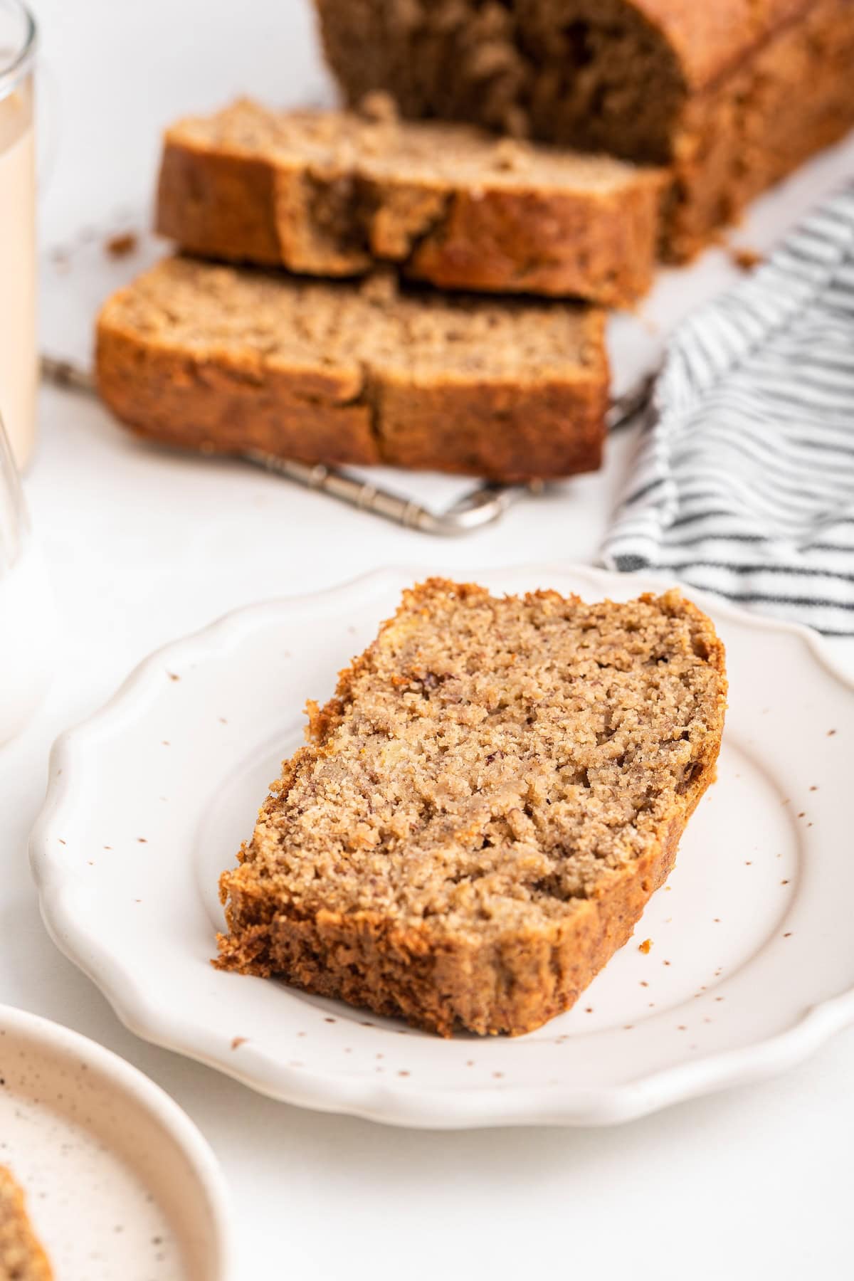 A slice of oat flour banana bread on a plate with the loaf of banana bread nearby.