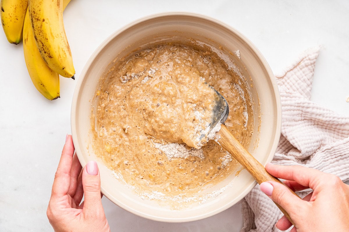 A woman's hand uses a silicone spatula to combine the wet and dry ingredients used for an oat flour banana bread in a large bowl.