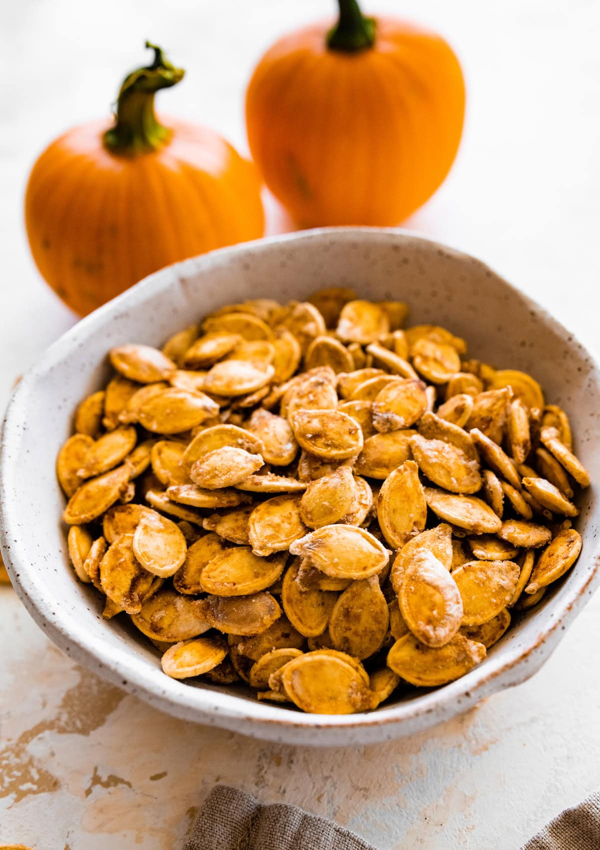 A bowl of roasted and salted pumpkin seeds near two small pumpkins.