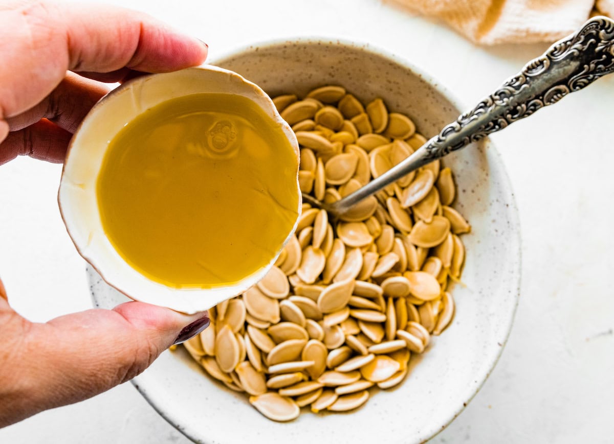 A womans hand pours olive oil into a bowl of pumpkin seeds.