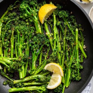 A pan with cooked and seasoned broccolini with fresh lemon wedges.