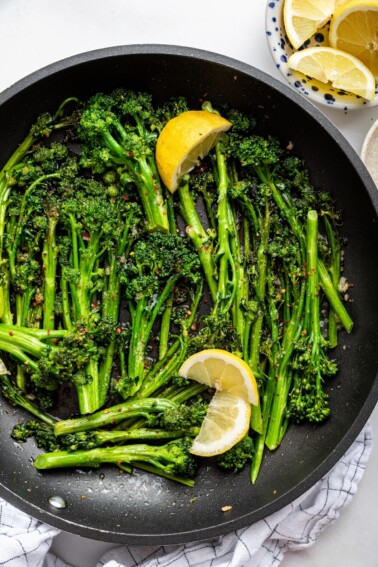 A pan with cooked and seasoned broccolini with fresh lemon wedges.