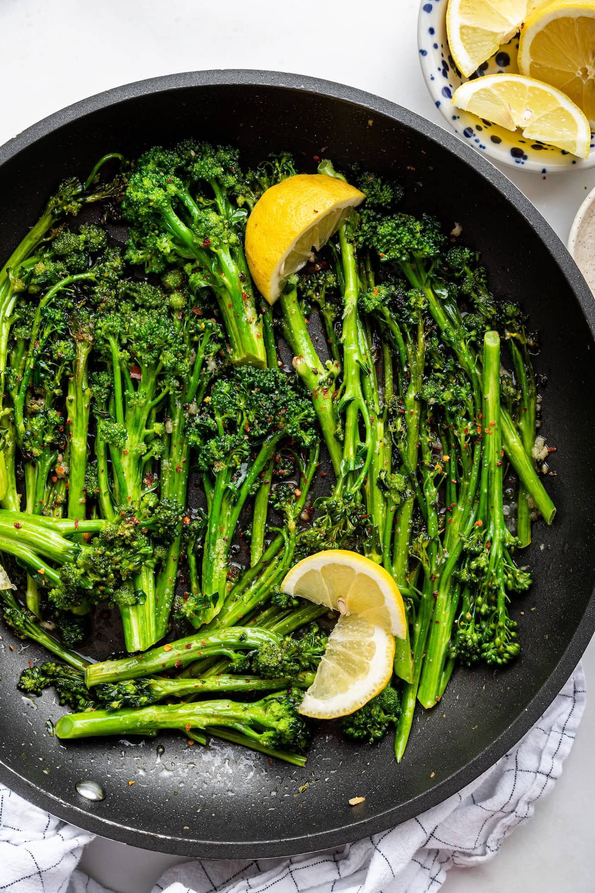 A pan with cooked and seasoned broccolini with fresh lemon wedges.
