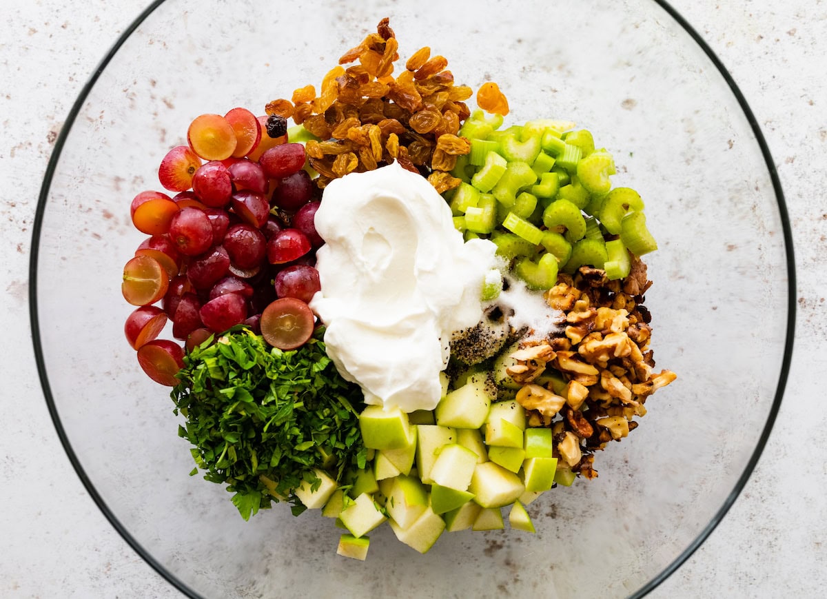 The ingredients for a waldorf salad in a glass bowl: grapes, golden raisins, celery, apples, pecans, parsley and yogurt.