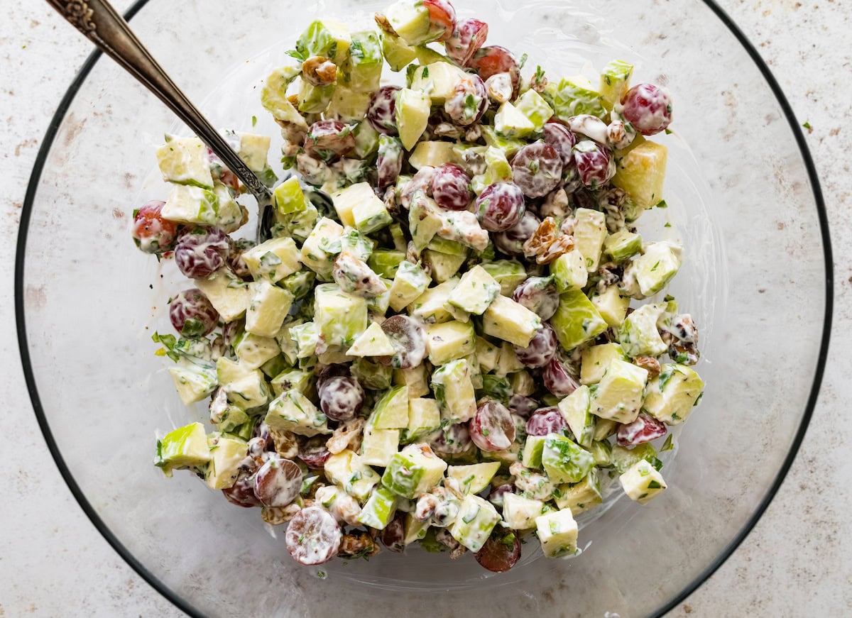 A mixed together waldorf salad with sliced apples, grapes, walnuts and more in a glass bowl.