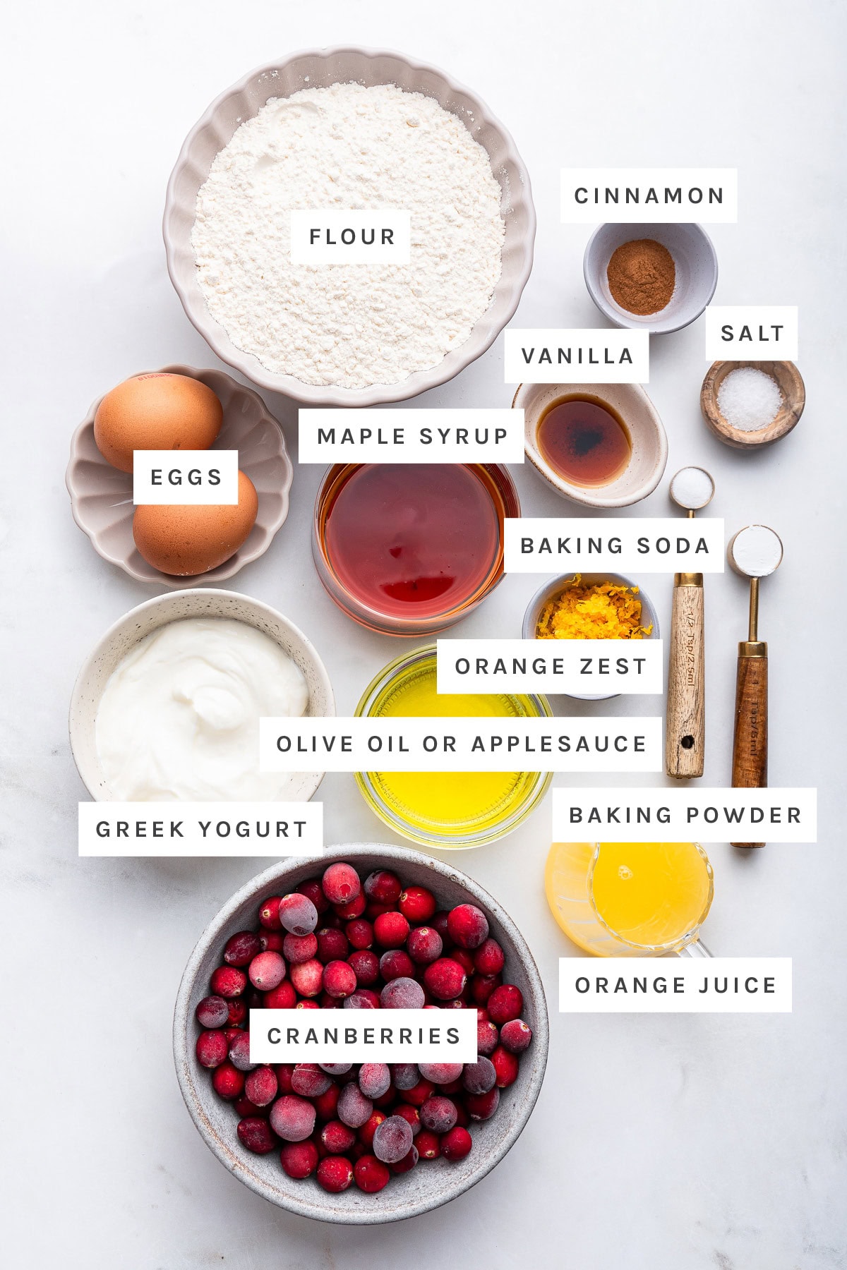 Ingredients measured out to make Cranberry Orange Muffins: flour, cinnamon, vanilla, salt, maple syrup, eggs, baking soda, orange zest, olive oil, Greek yogurt, baking powder, cranberries and orange juice.