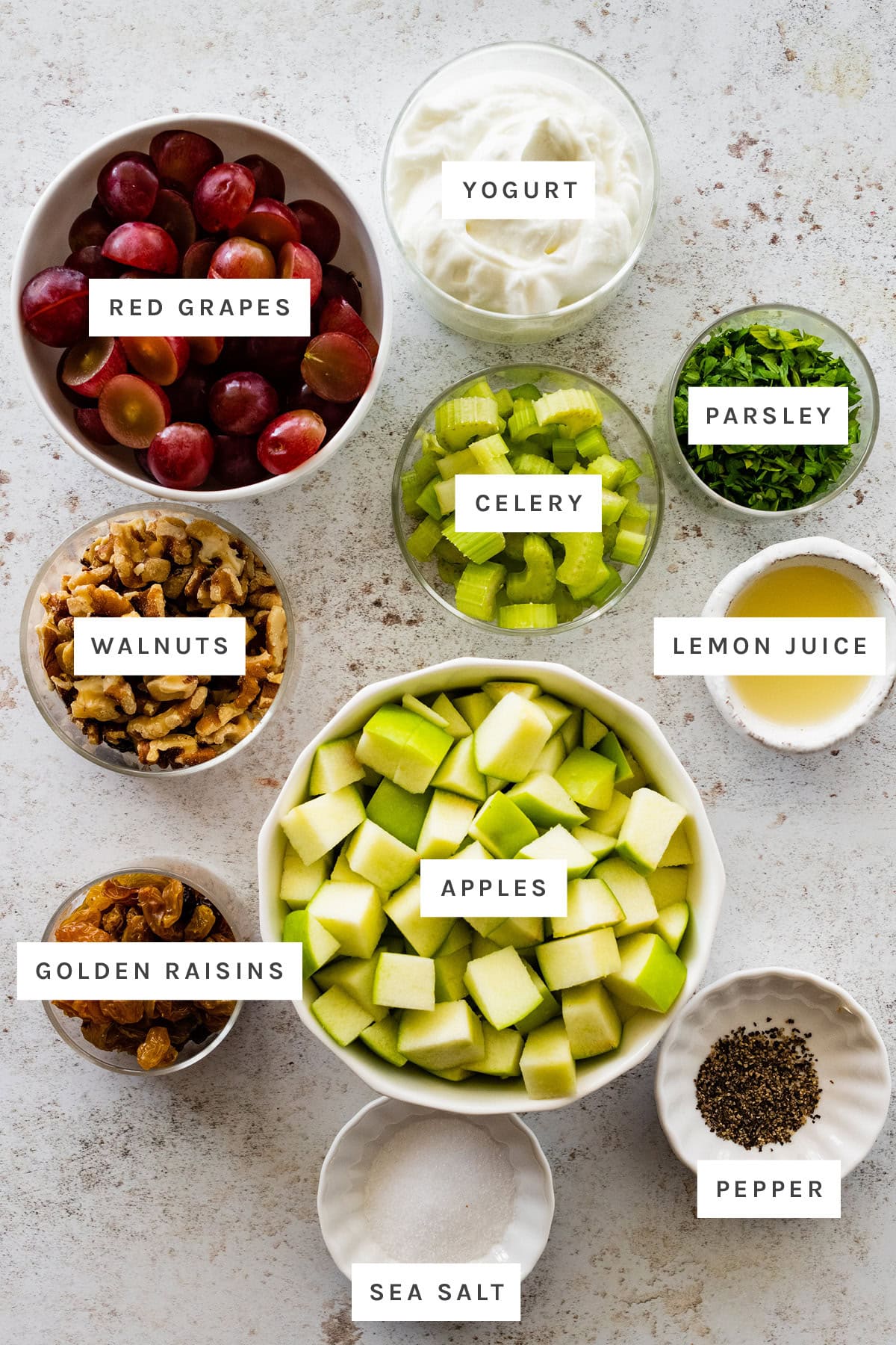 Ingredients measured out to make Waldorf Salad: red grapes, yogurt, parsley, celery, lemon juice, walnuts, apples, golden raisins, sea salt and pepper.