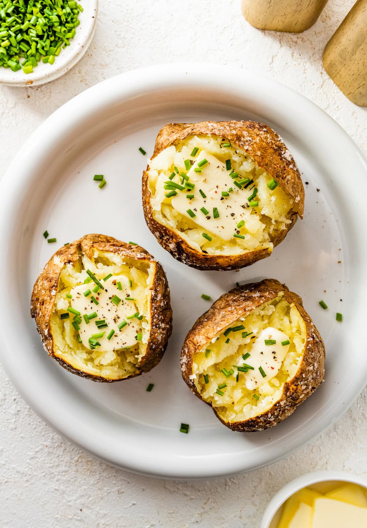 Three air fryer baked potatoes split open on a plate with slices of butter, seasoning, and fresh chives.