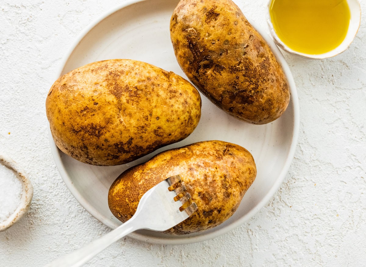 A fork poking a hole in a potato near two other potatoes on a plate.