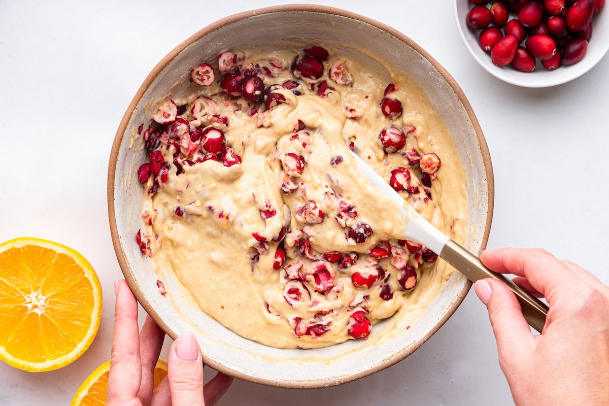 A woman's hands use a silicone spatula to combine cranberries in a muffin batter.