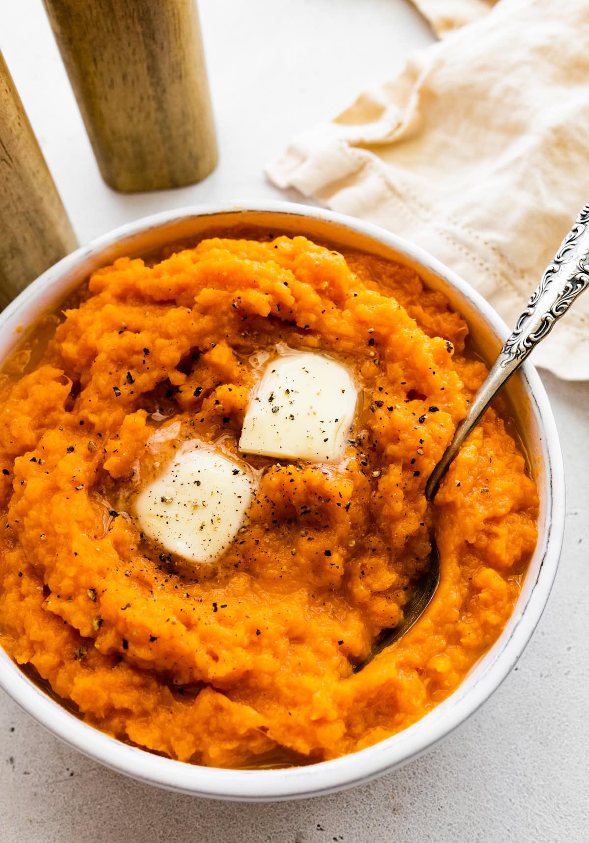 A white bowl of sweet mashed potato with black pepper, butter, and a metal spoon.