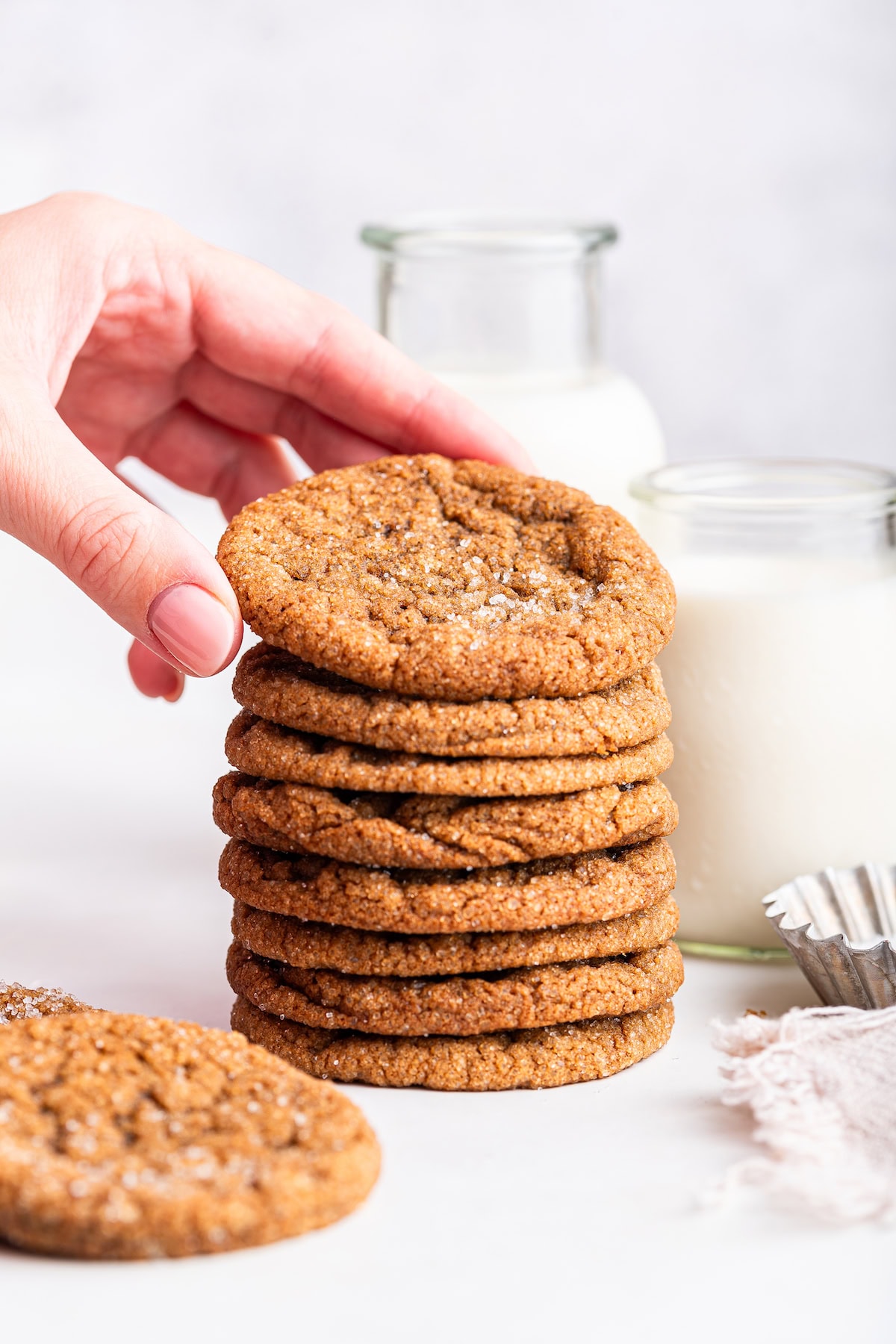 A woman's hand grabbing a molasses cookie from a stack of cookies. A glass of milk is in the background.