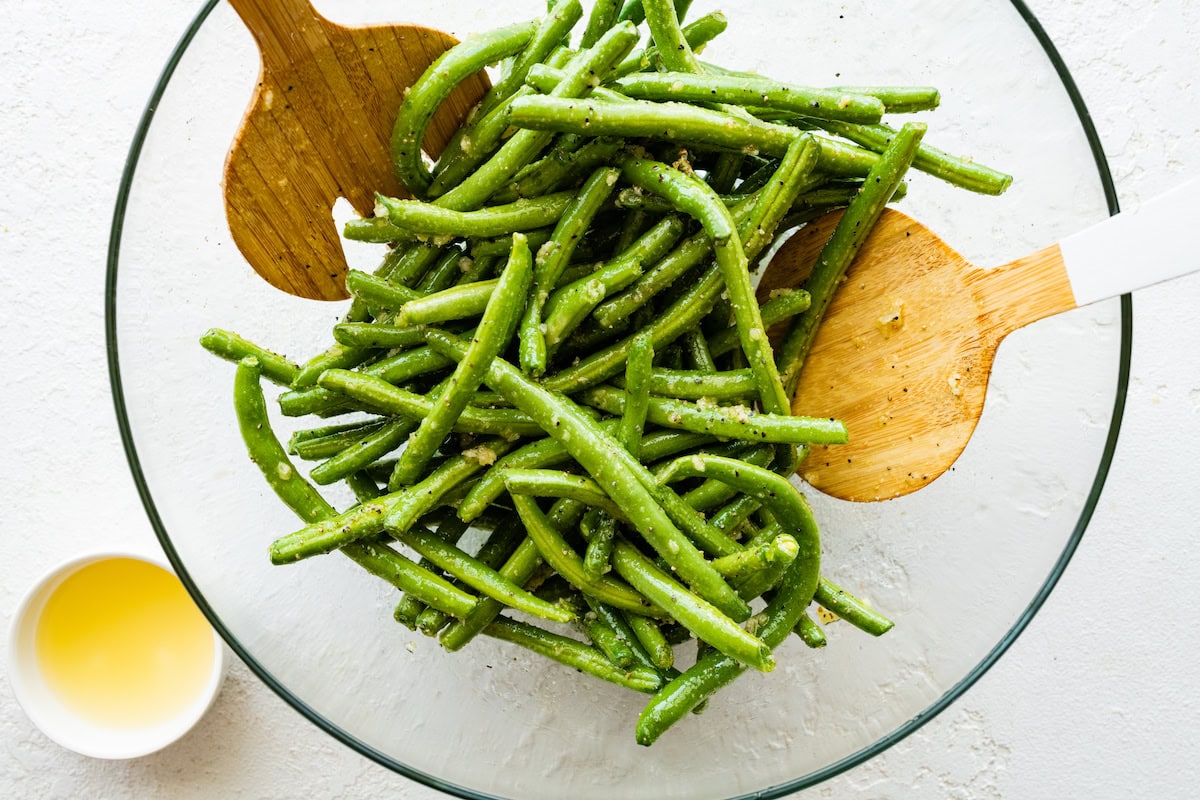 A large glass bowl of seasoned green beans with two wooden spatulas.