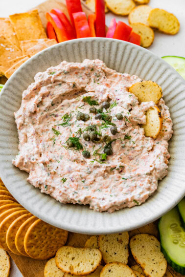 A bowl of smoked salmon dip, garnished with fresh dill and capers, served with crackers and fresh vegetables.