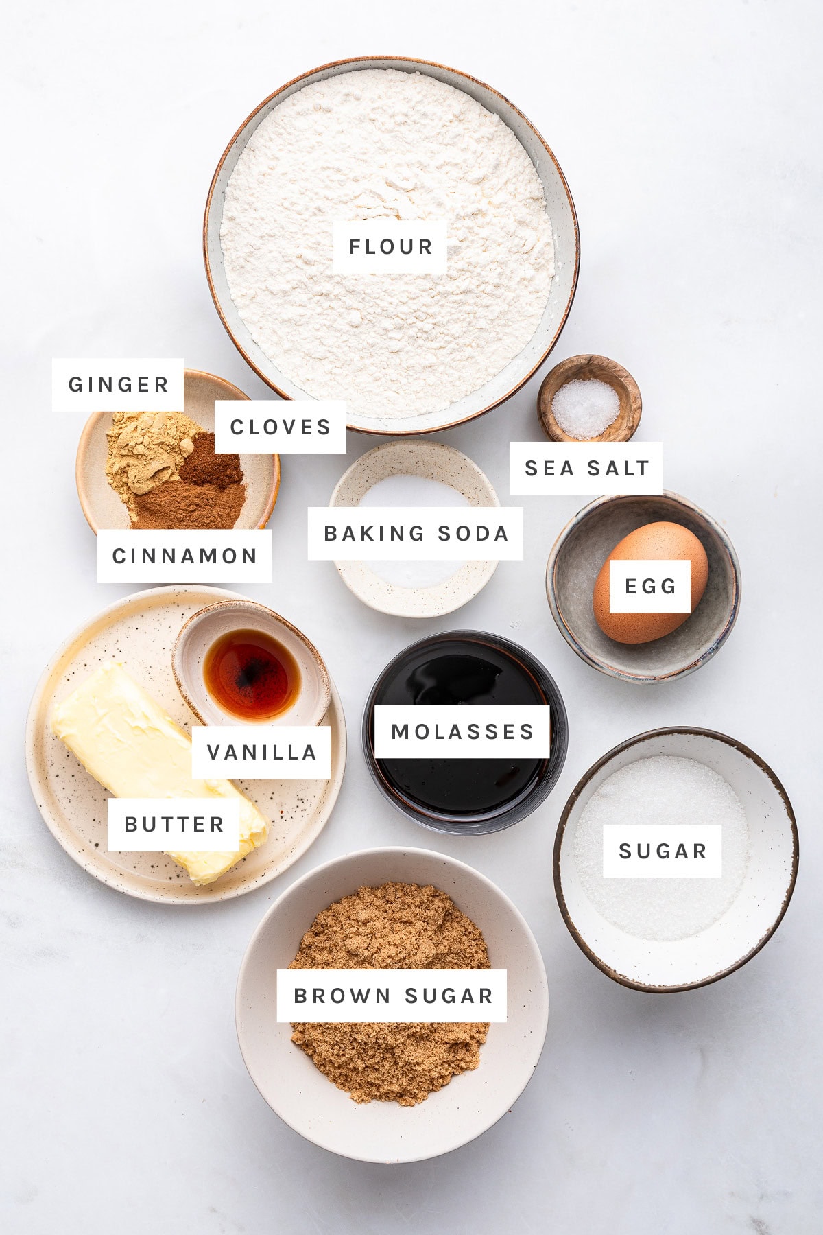 Ingredients measured out to make Molasses Cookies: flour, ginger, cloves, baking soda, sea salt, cinnamon, egg, molasses, vanilla, butter, brown sugar and sugar.