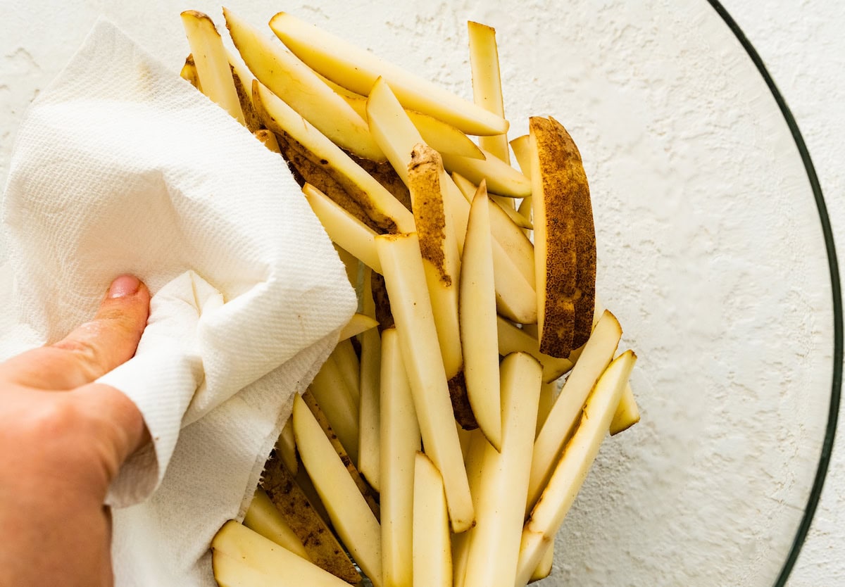 A woman's hand uses a paper towel to pat dry french fries in a large glass bowl.