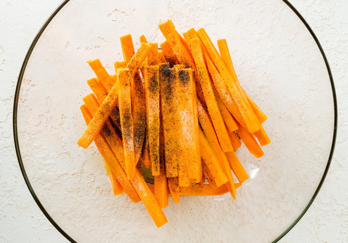 Sweet potato fries in a glass mixing bowl topped with oil, salt and pepper.