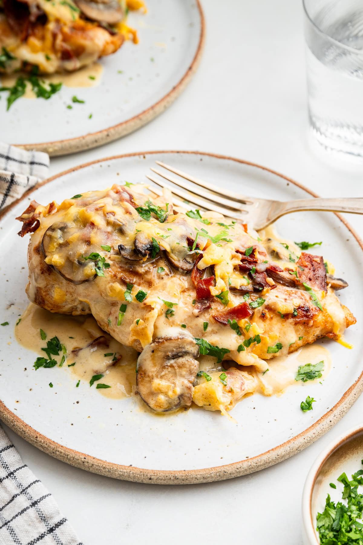 One chicken breast served on a white plate with a fork. The chicken breast is topped with turkey bacon, mushrooms and shredded cheese.