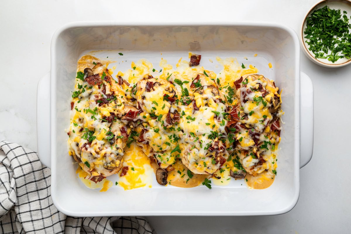 Four chicken breasts in a white baking dish topped with mushrooms, turkey bacon, melted cheese and fresh parsley.