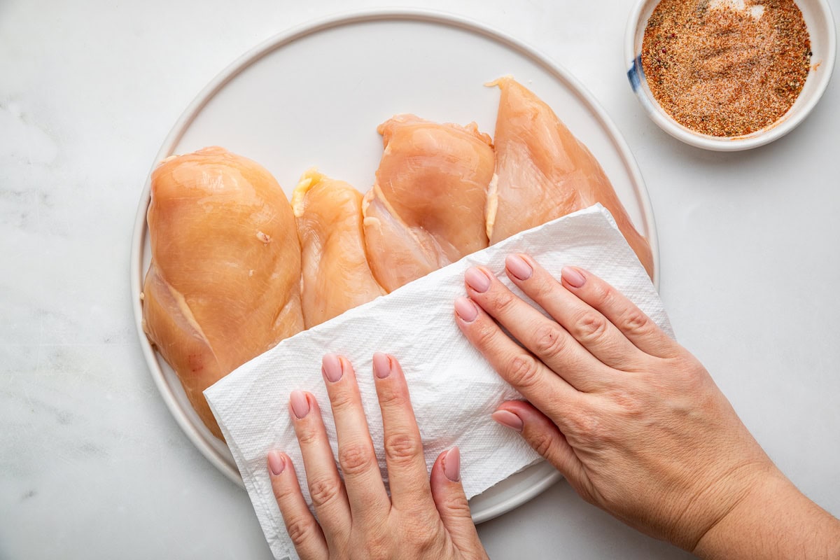 Woman's hands patting four chicken breasts dry with a paper towel.