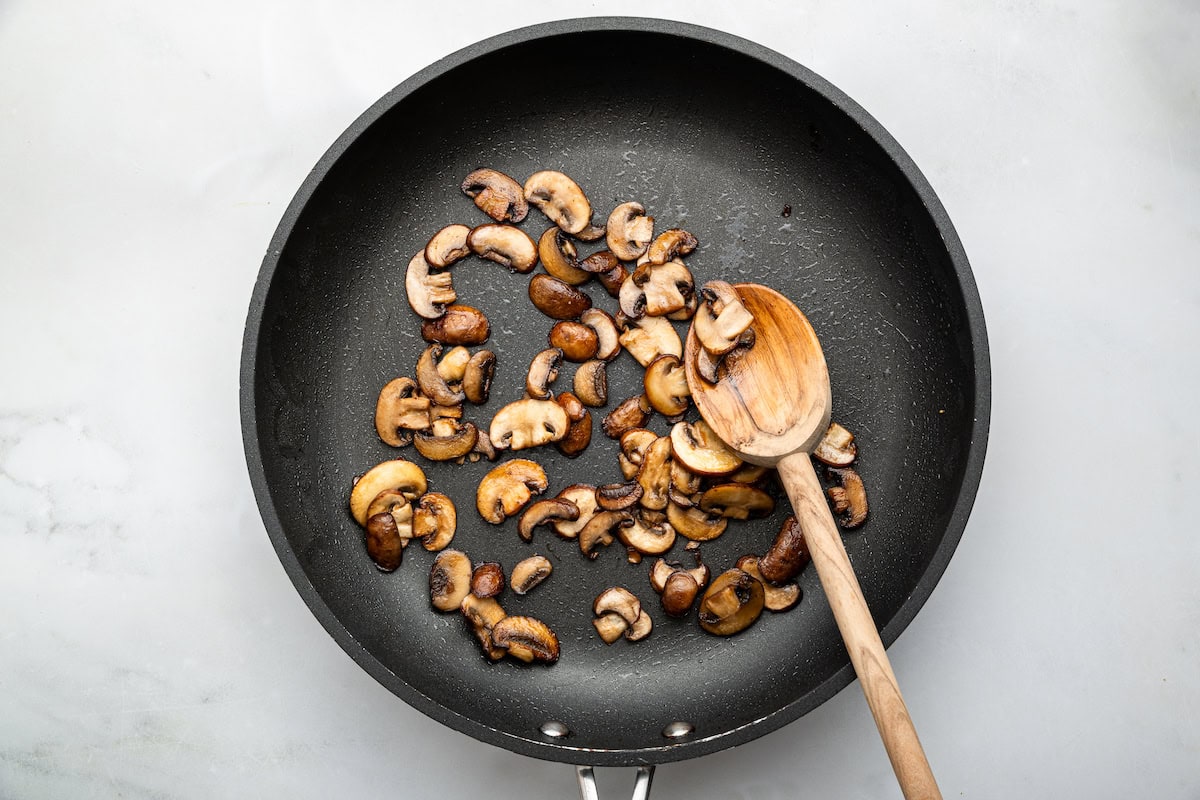 Sauteed msurhooms in a skillet with a wooden spoon.