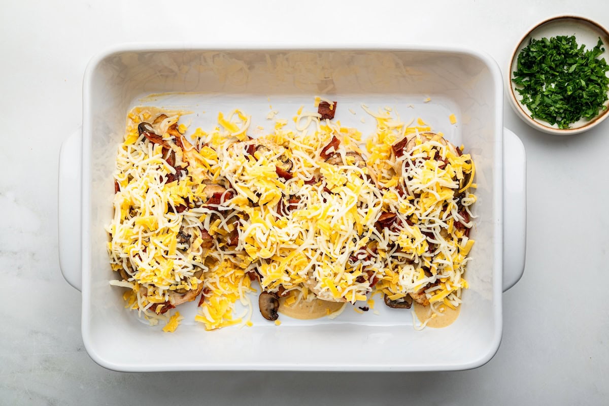 Four chicken breasts in a white baking dish topped with mushrooms, turkey bacon and shredded cheese.