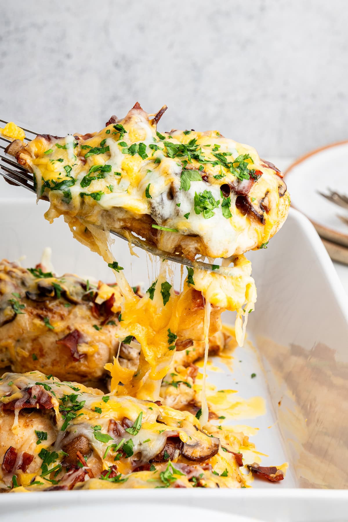 A chicken breast being served from a white baking dish. The chicken breast is topped with turkey bacon, mushrooms and shredded cheese.