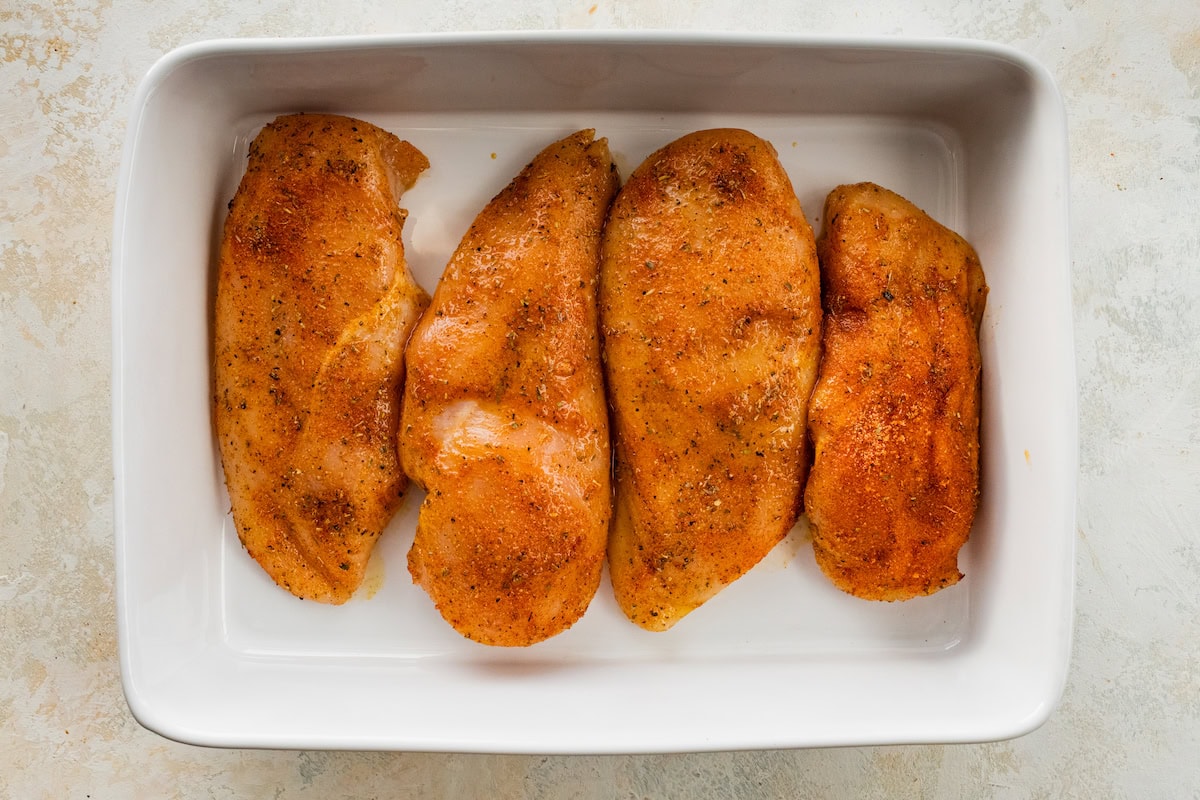 Four seasoned and raw chicken breasts in a large white baking dish.